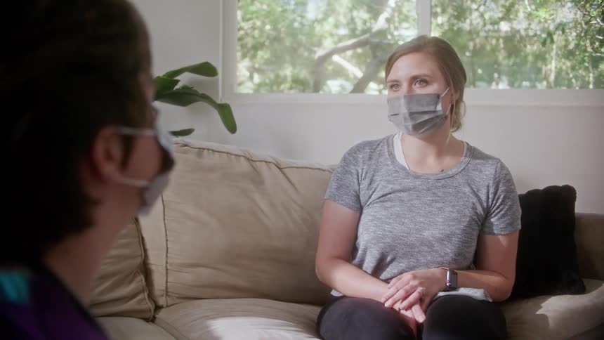 A nurse nods while listening to a patient talk as she sits on a couch at home