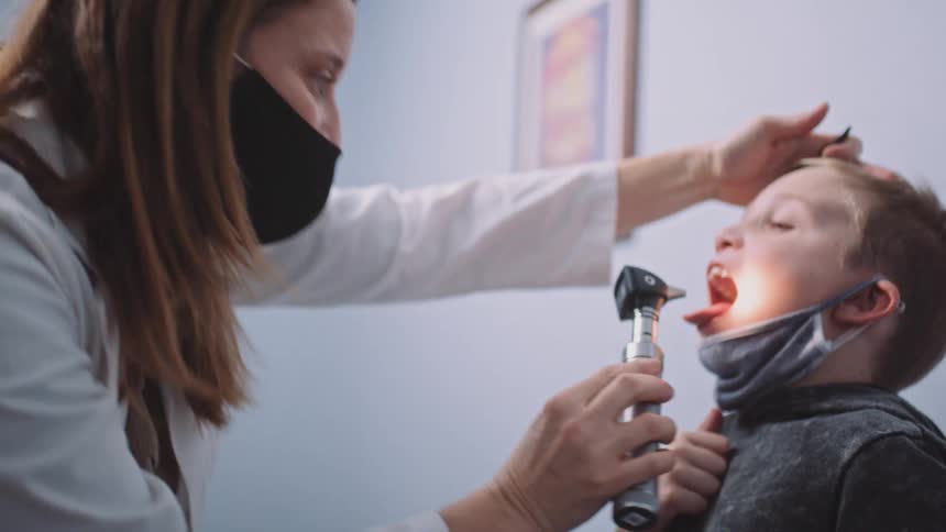 A female doctor uses an otoscope to examine the nose and mouth of a boy in a doctors office