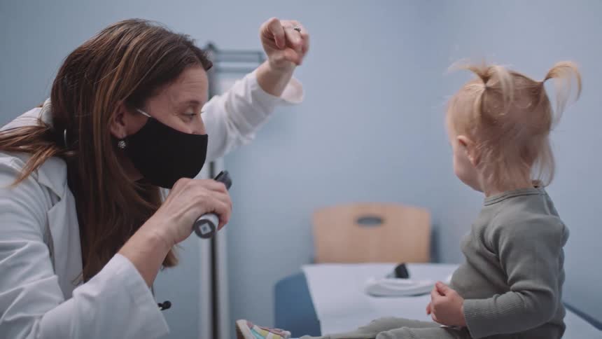 A female doctor using an ophthalmoscope to examine the eyes of a small girl snaps her fingers to get the child to face forward