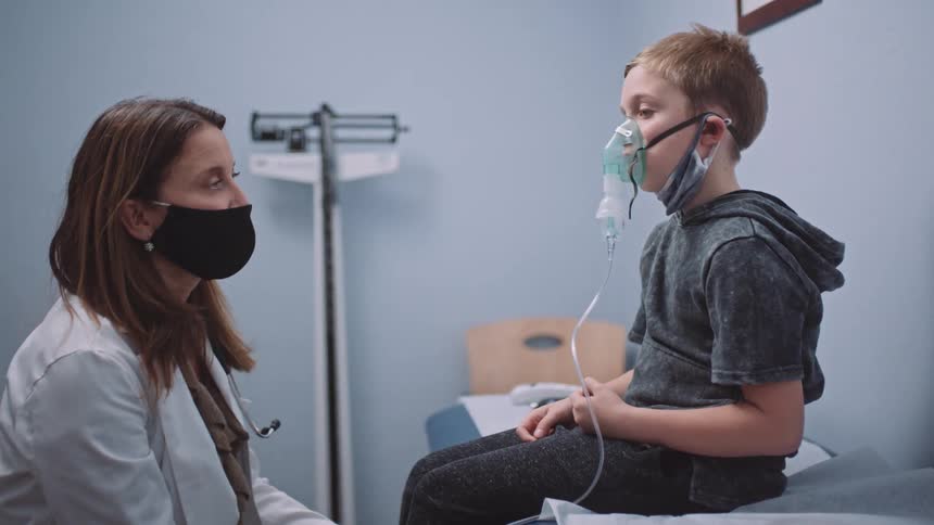 A female doctor in a doctors office speaks with a boy in a ventilation mask sitting on an examination table in a doctors office