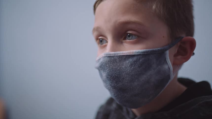 A young boy wearing a face mask sits and stares while waiting in a clinic exam room