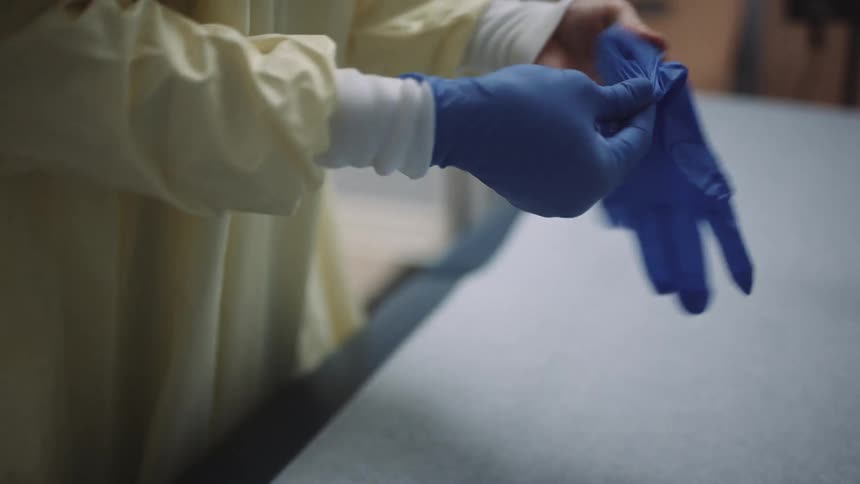 A doctor wearing a surgical apron puts on a pair of blue latex gloves while standing in a doctors office