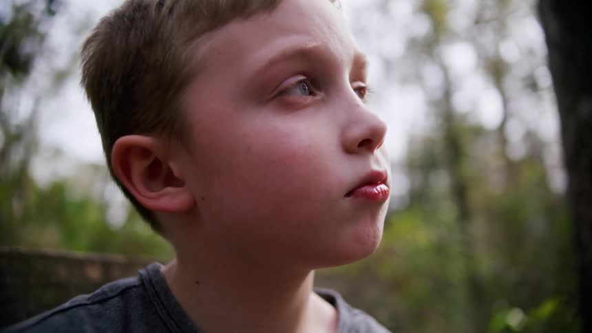 A young boy sits outside while surrounded by trees and stares off into the distance while thinking
