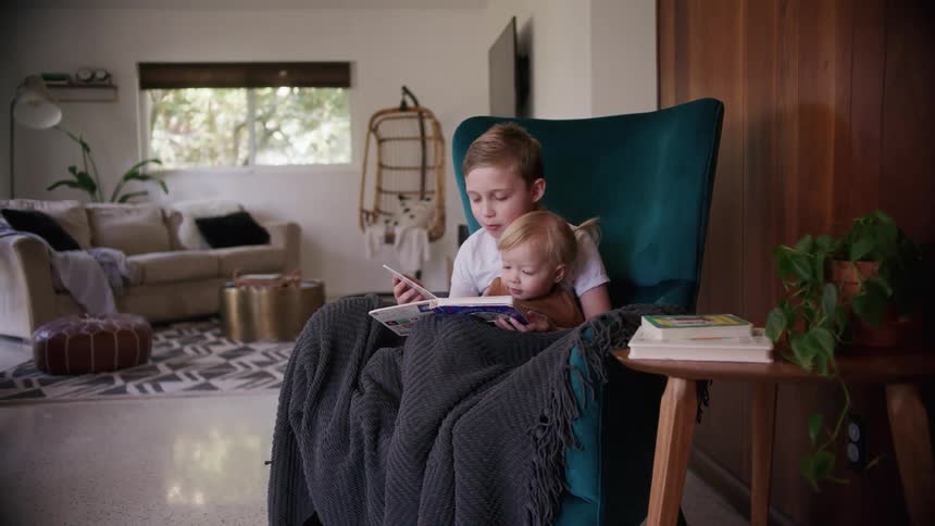 A young boy sits with his toddler sister and turns pages of her book while reading