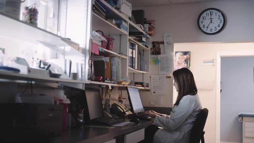 A woman in a lab coat sitting on a chair at a desk typing on a laptop