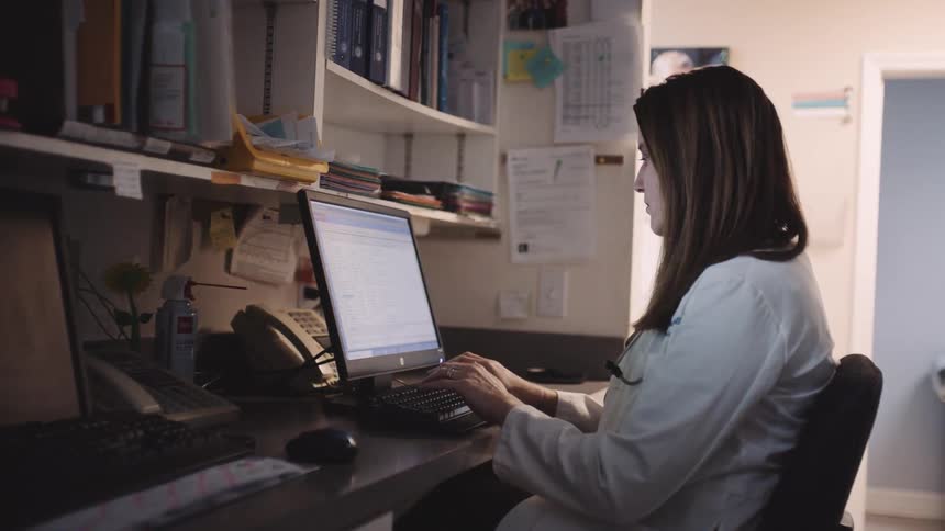 Doctor sits at desk entering information in computer across hall from examining room