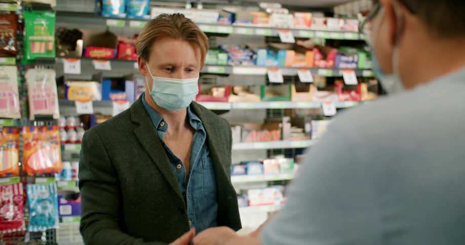 A man in a medical mask buying a pair of bananas in a store recieves a receipt from the masked cashier