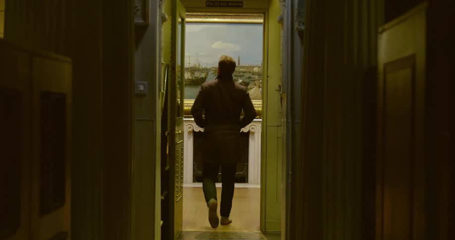 A man walks down a hallway with his hands in his jacket pockets and stops in front of a painting on a wall
