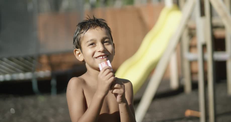 A young boy eats a snack while standing in his backyard next to a swing set