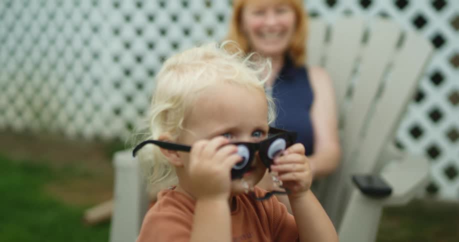 A child in a backyard putting on a toy glasses and mustache with a lady sitting on a chair in the background