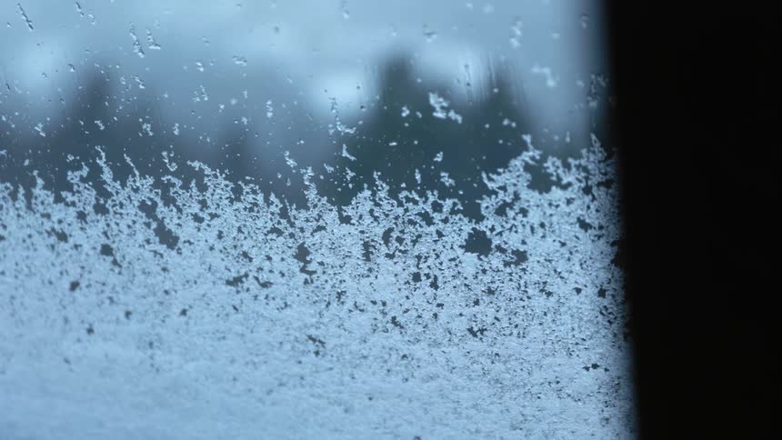 Snowflakes fall onto and slide down the surface of a window during the day