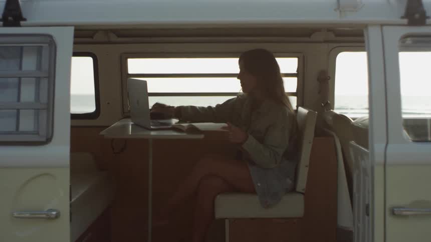 A young woman uses a laptop and writes in a notebook while sitting at a table in a large van parked on the coast during the day
