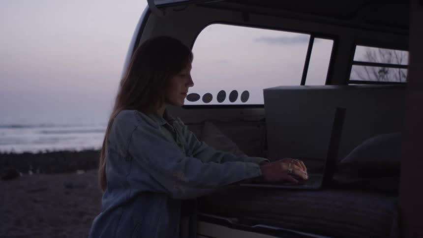 A woman typing on a laptop while standing next to an open van door on a beach