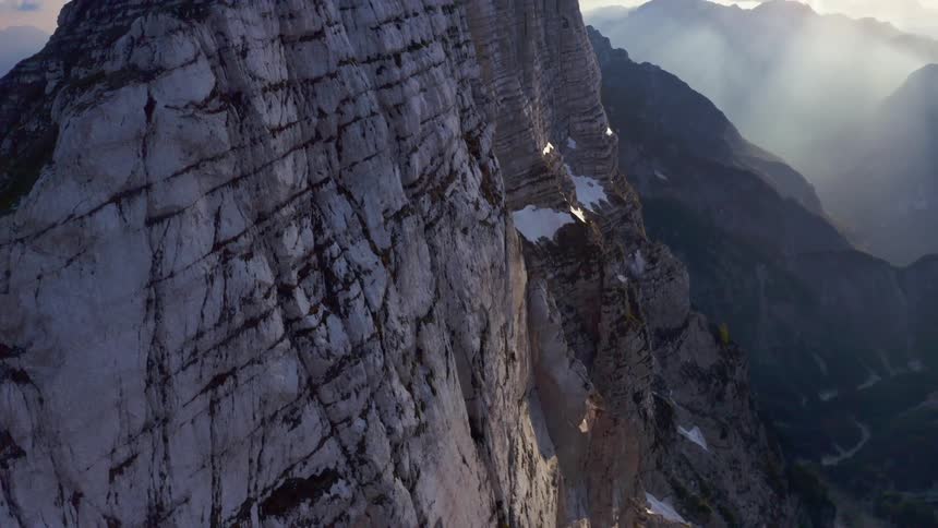 A sheer rocky mountain ridge dotted with snow with sunlight and peaks behind