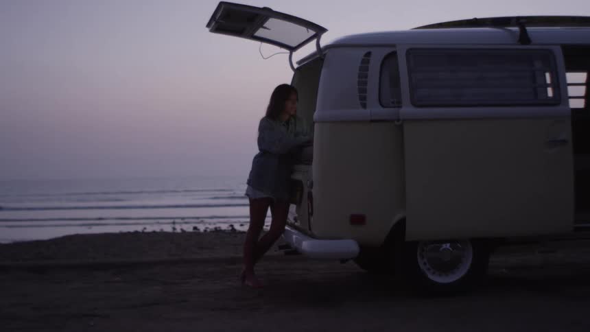 A young woman leans against the open rear end of a van parked on the coast on a cloudy evening