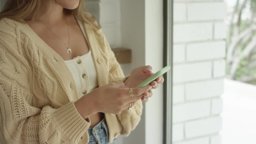 A woman standing in a room scrolling on a smart phone next to a window during the day