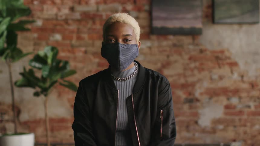 A young woman in a medical mask stares into the camera while sitting indoors