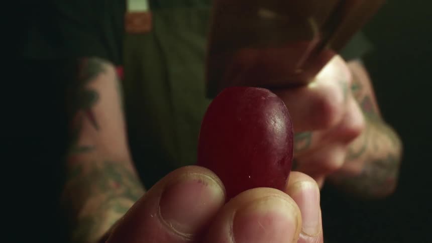 A man holds a red grape in his fingers and slices the top off with a sharp knife