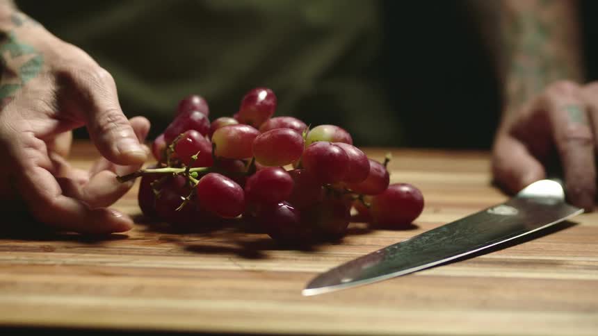 A man places a bunch of grapes beside a knife on a wooden cutting board