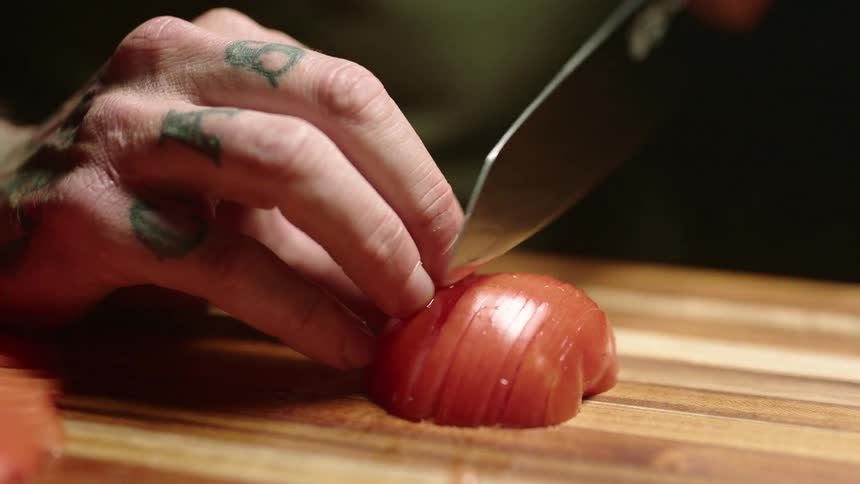 A person holds a tomato on a cutting board and carefully cuts thin slices it with a sharp knife