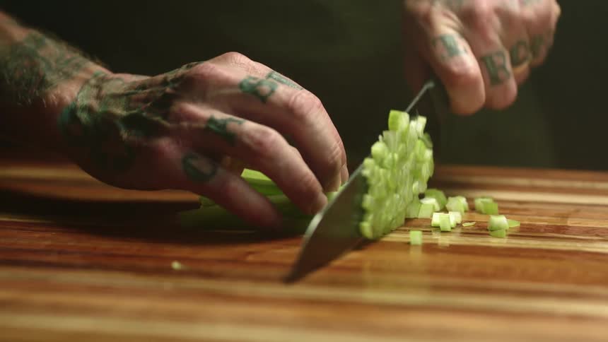 Close up of a tattoed man chopping celery with a sharp kitchen knife over a wooden board
