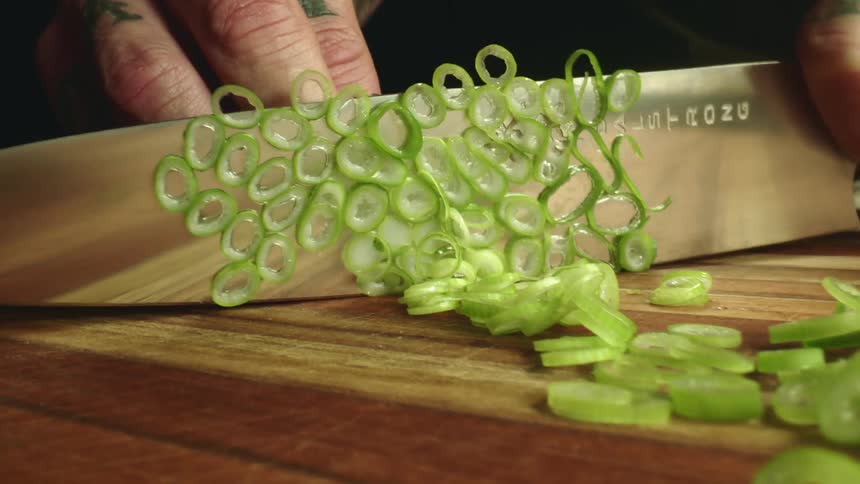 Close up of a tattoed man cutting chives with a sharp kitchen knife over a wooden board