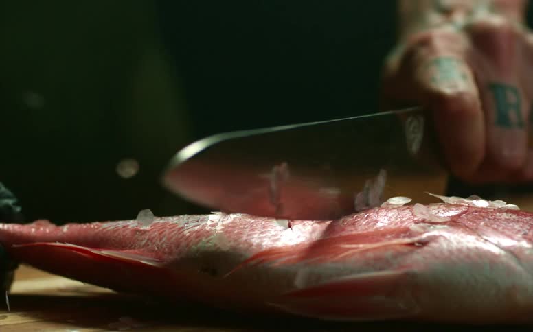 A man uses a knife to descale a fish on a cutting board