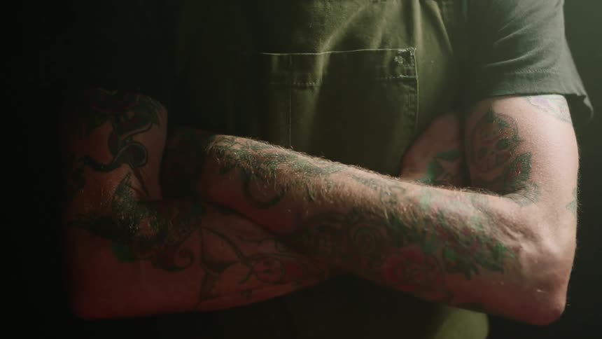 A heavily tattooed man stands and crosses his arms