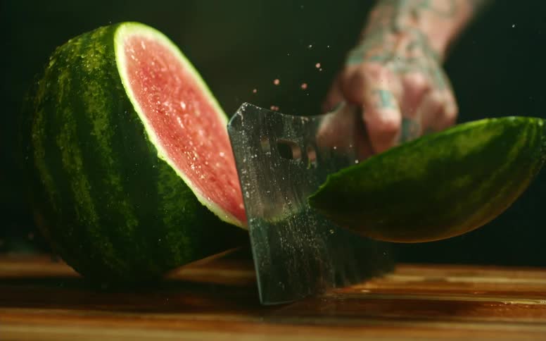 A tattoed man slicing through a watermelon using a sharp cleaver on a wooden cutting board