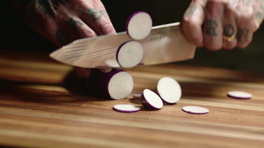 Close up of a tattoed man slicing eggplant with a sharp kitchen knife over a wooden board