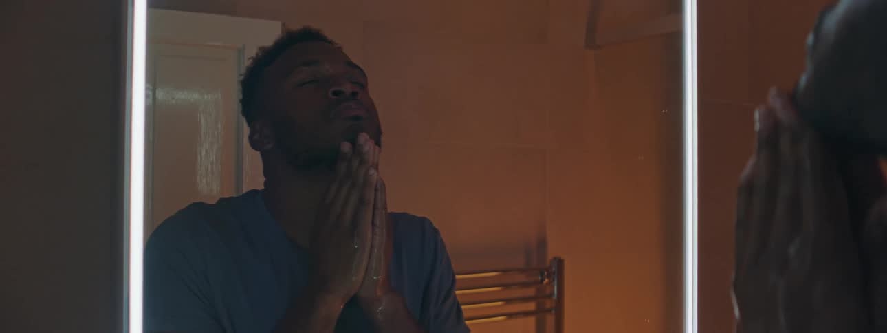 Young man stands in front of a mirror and rubs his face with his hands before washing
