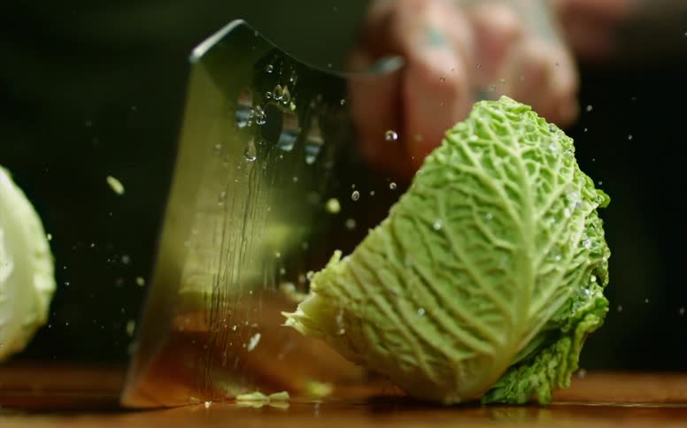 Close up of a tattoed man chopping a cabbage with a sharp cleaver over a wooden board