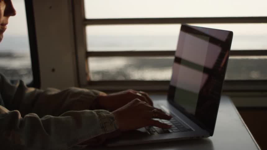 A woman types on her laptop while sitting in the back of her van parked near the ocean