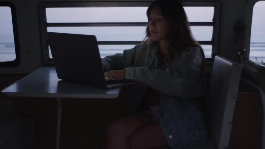 A woman in a vehicle sitting on a chair next to a table typing on a laptop