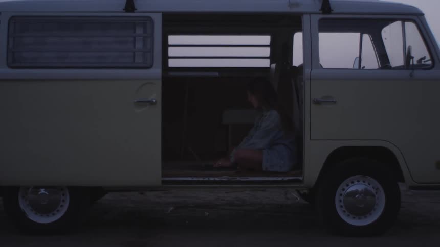 A young woman types on a laptop while sitting on the floor of a large van parked offroad on a cloudy evening