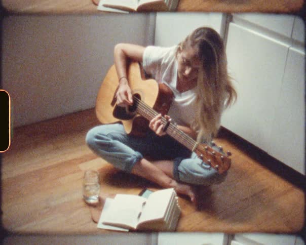 A vintage film with a teenage girl sitting on the floor at home playing the guitar and writing in a book