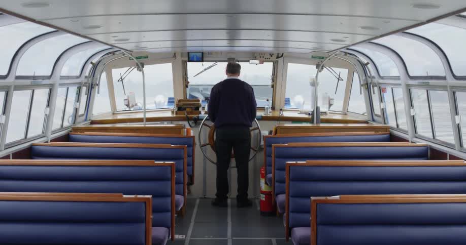 A man stands at the ships wheel with empty bences behind him on a ferry