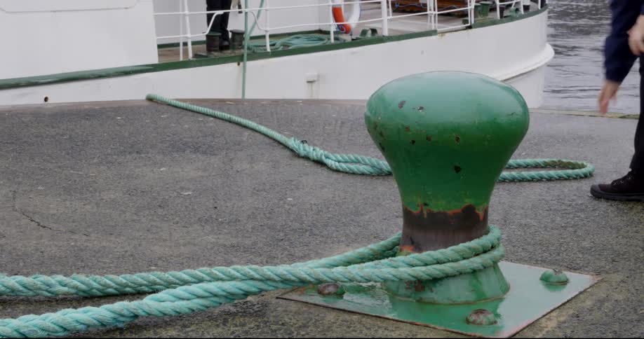 A sailor on a boat tosses one end of a docking line to the dock where another person retrieves it and places it around the docking cleat