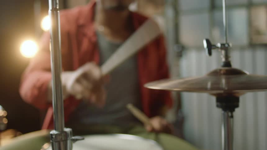 A man sitting and playing the drums with drumsticks with a wall in the background