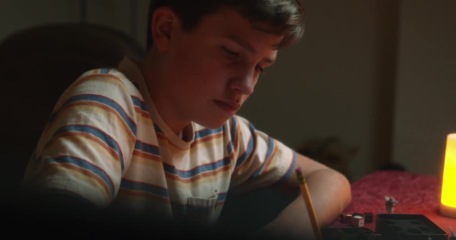 A young man sits at a wooden desk and writes using a standard wooden pencil