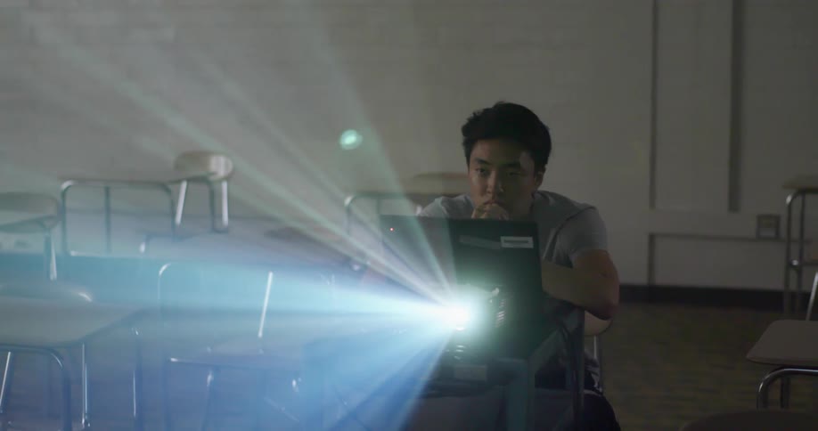 A young asian man sitting at a desk in a classroom watches a projection from a projector attached to a laptop on the desk in front of him