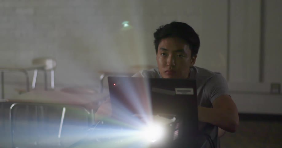 A boy sits at a school desk with an open laptop in front of him and a glaring light from a projector