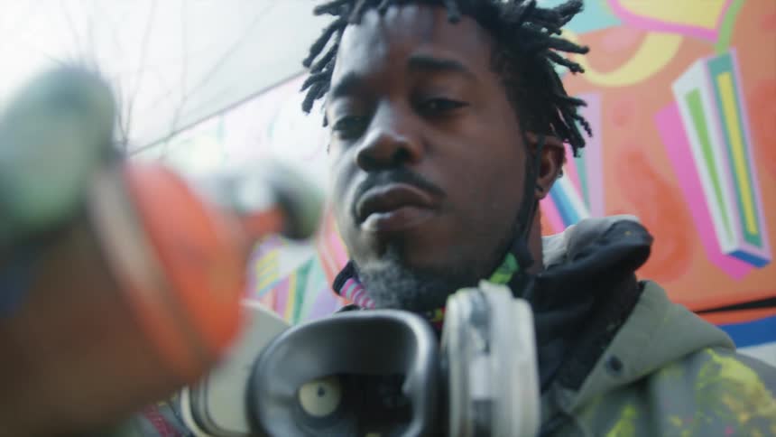 A painter shakes a can of spray paint and points the nozzle towards the camera for a moment before laughing and moving it away
