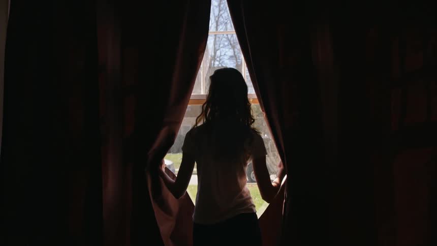 A girl stands at a window and parts the curtains to look outside