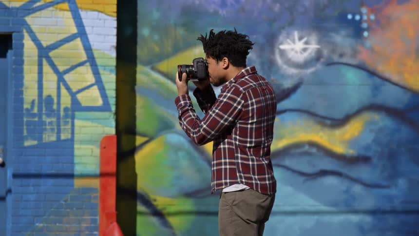 A man takes a photo with a camera while standing next to a graffiti wall