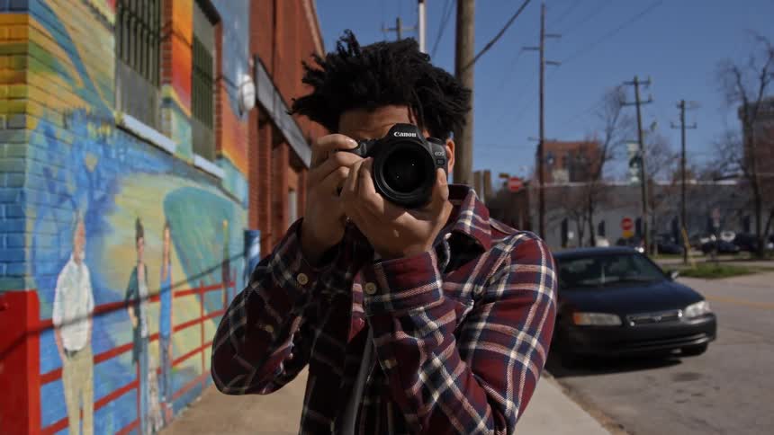 A man stands on a sidewalk near a mural and raises a camera to his eyes