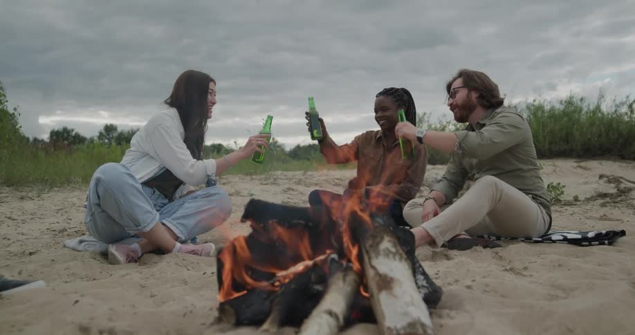 Friends sits on beach near campfire drinking under overcast sky