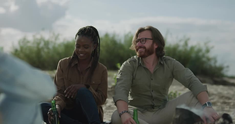 A mixed group of people hold alcohol and laugh around a campfire on a beach