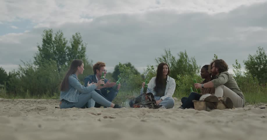 Friends sit at beach around campfire drinking