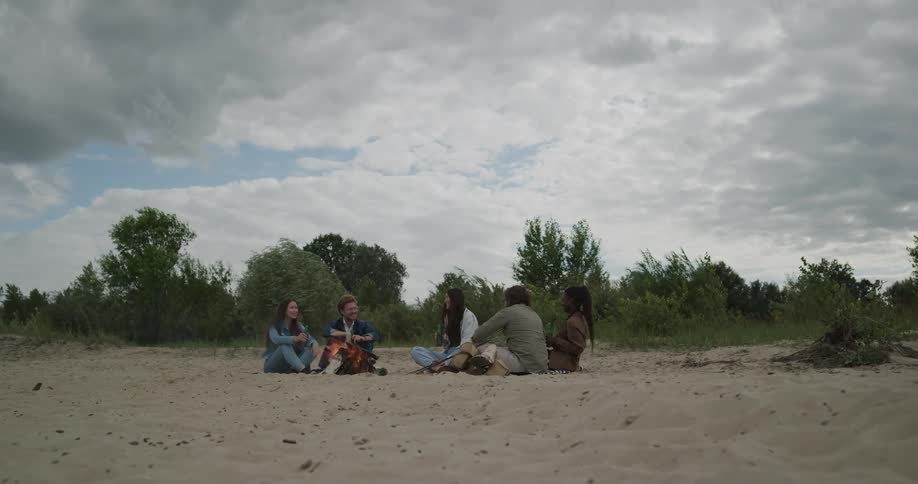 A group of friends sit around a campfire on a beach and talk and laugh